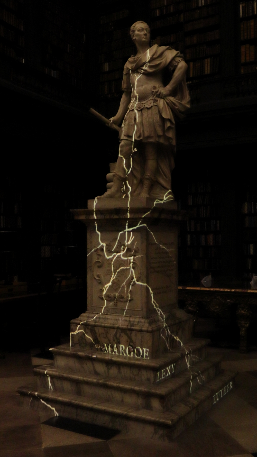 Marble statue of Codrington dressed in Roman style, on a tall plinth. Cracks are projected onto the statue and plinth, along with the names "Margoe", "Lexy", and "Luther".